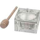 A square clear crystal honey dish with an etched design on its side, a matching lid, and a wooden honey dipper resting next to it.