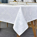 A white tablecloth with a woven damask pattern covering a wooden dining table. The tablecloth's corner is folded up to show the texture. The table is set with plates with gold trim and wine glasses.