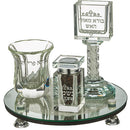 An elegant crystal Havdalah set on a mirrored base with four crystal feet. The set includes a twisted crystal candle holder, a tall goblet, and a spice box, all featuring engraved Hebrew text.