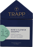 A lighted, white scented candle in a clear glass votive, positioned next to its dark blue, house-shaped packaging box. The box features the "TRAPP FRAGRANCES" logo in gold, and a label indicating "No. 13 BOB'S FLOWER SHOPPE™" with scent notes of "FRESH FLORALS, CUT GREEN STEMS, WATERY ACCORDS." The candle is a "FRAGRANCED CANDLE 7 oz | 198g ℮".