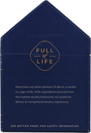 A lighted, white scented candle in a clear glass votive, positioned next to its dark blue, house-shaped packaging box. The box features the "TRAPP FRAGRANCES" logo in gold, and a label indicating "No. 13 BOB'S FLOWER SHOPPE™" with scent notes of "FRESH FLORALS, CUT GREEN STEMS, WATERY ACCORDS." The candle is a "FRAGRANCED CANDLE 7 oz | 198g ℮".