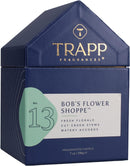 A lighted, white scented candle in a clear glass votive, positioned next to its dark blue, house-shaped packaging box. The box features the "TRAPP FRAGRANCES" logo in gold, and a label indicating "No. 13 BOB'S FLOWER SHOPPE™" with scent notes of "FRESH FLORALS, CUT GREEN STEMS, WATERY ACCORDS." The candle is a "FRAGRANCED CANDLE 7 oz | 198g ℮".