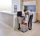 A two-tiered stainless steel trash and recycling bin with a black lid and base. The top compartment has a small, horizontal orange light on its front.