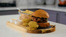 Yukon Glory Burger Serving Set, featuring a wooden board with a metal fry basket, two ceramic ramekins for sauces, and a burger on a round black coaster.