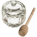 A small, faceted crystal honey pot with a matching lid sits next to a wooden honey dipper. Hebrew writing is visible on the lid of the honey pot.