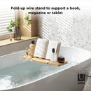 A light wood Umbra bathtub caddy with expandable metal arms, featuring a slotted book/tablet stand, a wine glass holder, and a recessed area with drain holes for soap or accessories.