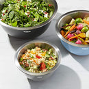 Three OXO Good Grips stainless steel mixing bowls nested inside each other, showing their gray, light blue, and dark gray exteriors and shiny stainless steel interiors. Each bowl has a white OXO logo and appears to have a non-slip base.