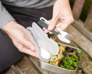 A stainless steel knife, fork, and spoon with black and silver handles, next to a translucent white rigid case. The handles are designed to fit together for compact storage.