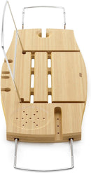A light wood Umbra bathtub caddy with expandable metal arms, featuring a slotted book/tablet stand, a wine glass holder, and a recessed area with drain holes for soap or accessories.