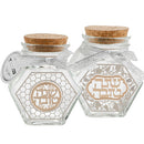 Two hexagonal glass honey jars with cork lids, tied with a sheer silver ribbon. The jar on the left has a gold honeycomb pattern and Hebrew text in a circular design. The jar on the right features a silver floral background with gold Hebrew text in an oval frame.