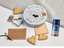 A large, themed gift set including a white tin printed with a chocolate chip cookie recipe, two cookies tied with a red bow, a blue can of Bartenura Moscato, and a small gold mask.