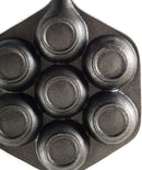 The image displays a cast iron aebleskiver pan. It's a dark-colored pan with a single handle and seven hemispherical indentations, which are used to cook aebleskiver (Danish pancake balls). The shape of the pan, excluding the handle, is hexagonal.