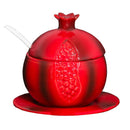 A red ceramic honey dish shaped like a pomegranate, with a matching lid and saucer. A small white ceramic spoon is resting in a notch on the lid.