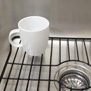 Two rectangular sink protector grids in a black finish, designed to fit in a kitchen sink, each with a central oval cutout for the drain and slightly raised feet, shown side-by-side.