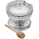 A clear, elegant crystal honey dish with a faceted design and a matching lid with Hebrew text. A wooden honey dipper rests in front of the dish.