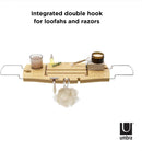 A light wood Umbra bathtub caddy with expandable metal arms, featuring a slotted book/tablet stand, a wine glass holder, and a recessed area with drain holes for soap or accessories.