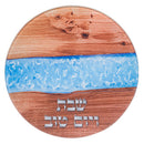 A circular wooden challah board featuring a prominent, wavy blue resin inlay running horizontally through its center. The top and bottom sections of the board display natural wood grain, and the Hebrew words "שבת" (Shabbat) and "ויום טוב" (Ve'Yom Tov - And a good day) are embossed or engraved in silver on the lower portion of the wood.