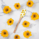 Tovolo Sunflower Field spatula with a wooden handle and sunflower design on the silicone head, shown on a marble counter, surrounded by sunflowers