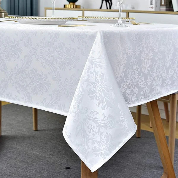 A white tablecloth with a woven damask pattern covering a wooden dining table. The tablecloth's corner is folded up to show the texture. The table is set with plates with gold trim and wine glasses.

