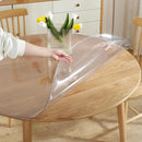 A clear, round vinyl table protector being laid flat on a light wood dining table.