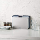A set of three rectangular cutting boards in gray and white, stored vertically in a silver stand. The boards feature metal handles and non-slip feet.