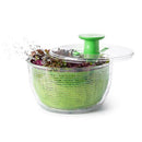 An OXO Good Grips Salad Spinner in action, showing wet, leafy greens inside a clear outer bowl and a green inner spin basket. A clear lid with a green pump handle is partially open, and droplets of water are flying off the spinning lettuce.