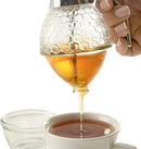Honey being poured from the Norpro glass dispenser into a steaming cup of tea, with the thumb lever pressed down and golden honey flowing smoothly.