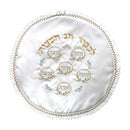 A round, white matzah cover with intricate embroidery. It has gold and silver threads showing Hebrew text and six small circles with more Hebrew words. The edge is decorated with a ruffled gold and silver trim.