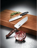 A Victorinox Santoku knife displayed alongside another Victorinox knife on a wooden cutting board with fresh herbs.