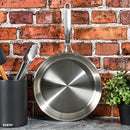 Babish stainless steel frying pan hanging on a kitchen wall against a brick backsplash.