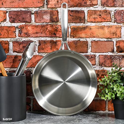 Babish stainless steel frying pan hanging on a kitchen wall against a brick backsplash.