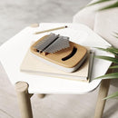 An elevated, side-angle view of an Umbra Strumba Kalimba. This musical instrument features a light-colored wooden top with a crescent-shaped sound hole and a series of white-tipped metal tines. The tines are engraved with musical notes or numbers. The base of the instrument is a smooth, rounded, off-white or cream color. The image is set against a plain white background. The "U umbra" brand logo is visible in the bottom right corner.