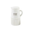 A white ceramic shaker cleverly shaped like a miniature pitcher. The design features a beaded top rim, vertical ribbing on the bottom half, and the black text 'Shake it out' on the side.