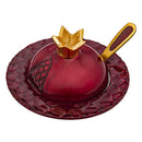 A vibrant red, pomegranate-shaped honey dish with a textured surface, resting on a matching round tray. A golden spoon with a red handle rests alongside the dish, which features a golden crown-like top.