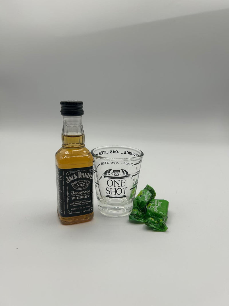 A mini bottle of Jack Daniel’s Tennessee Whiskey (50ml), a Fox Run 1.5 oz Measuring Shot Glass, and two Sour Green Apple Candies placed neatly on a white background.