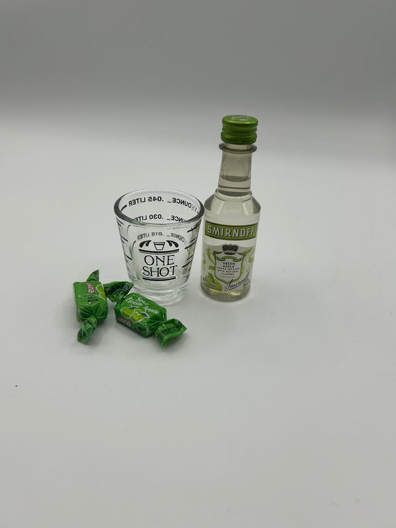 A mini bottle of Smirnoff Green Apple Vodka (50ml), a Fox Run 1.5 oz Measuring Shot Glass, and two sour green apple candies arranged on a clean white background.