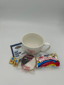 A gift set featuring a large white mug, a packet of hot cocoa mix, a black and white cookie, two small chocolate squares, and a bag of mini marshmallows.