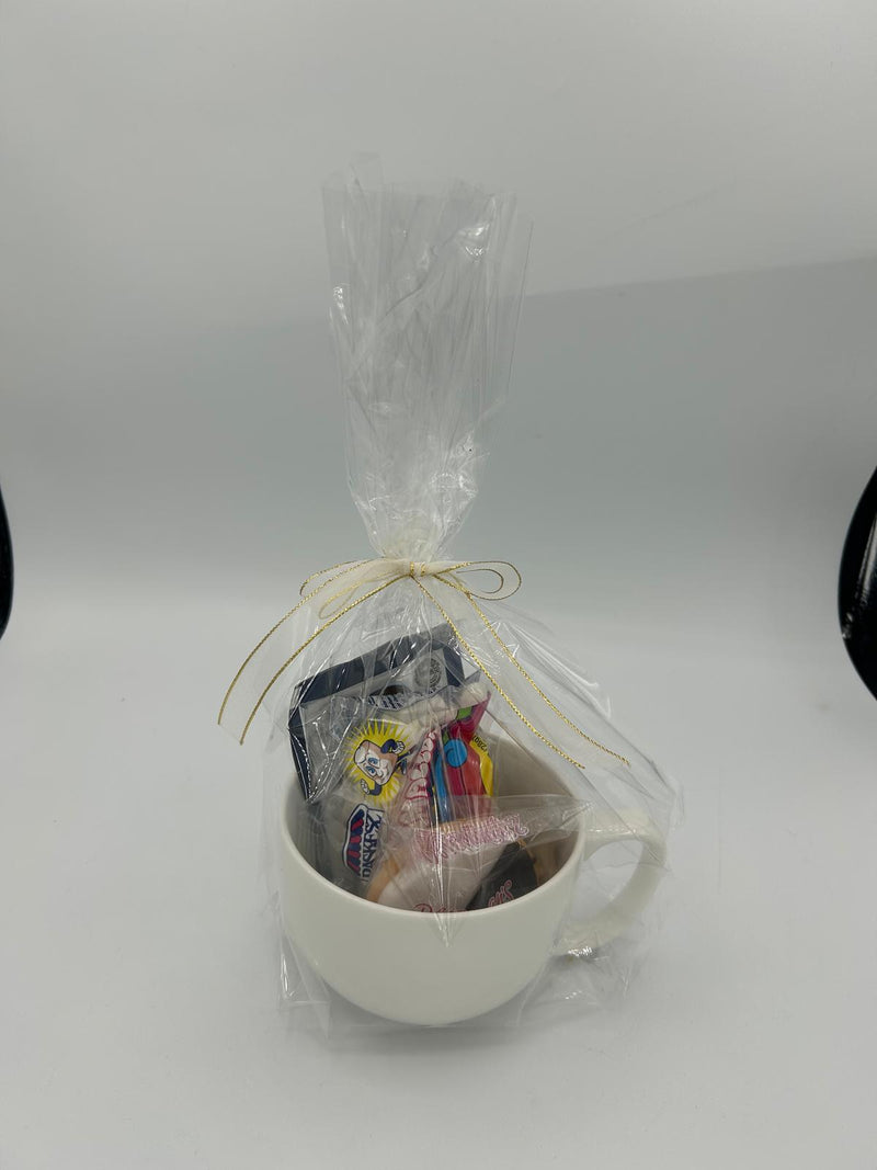 The Cozy Cocoa gift set wrapped in clear cellophane and tied with a gold ribbon, with the white mug and assorted treats visible inside.