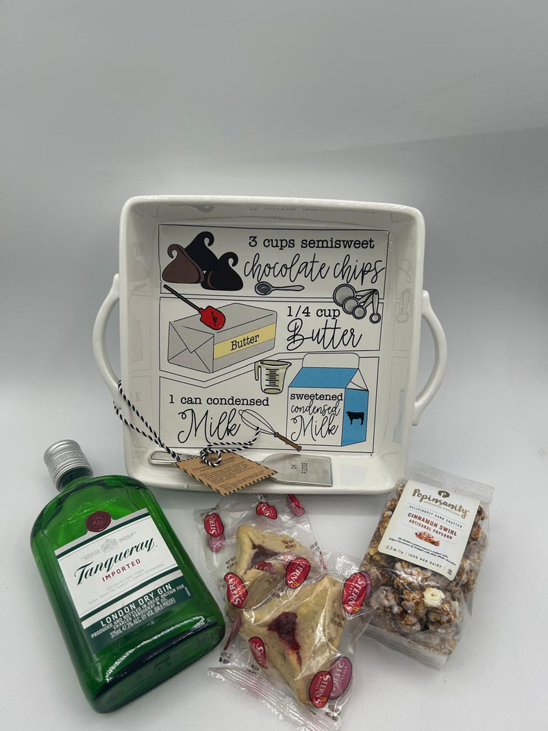 A close-up of a Shalach Manos gift set featuring a white ceramic fudge baking dish with built-in handles, a bottle of Tanqueray London Dry Gin, a bag of Popinsanity Cinnamon Swirl Popcorn, and two individually wrapped Stern’s Hamantaschen cookies. The dish has an illustrated fudge recipe on the inside, making it both functional and decorative.