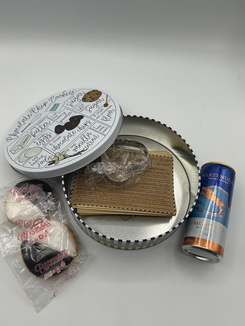 A close-up of an opened Purim gift tin, showcasing its contents: a metal cookie cutter, printed recipe cards, two Reisman’s Black and White Cookies, and a 375ml can of Bartenura Moscato. The tin’s lid features a charming chocolate chip cookie recipe illustration, adding a unique and festive touch.