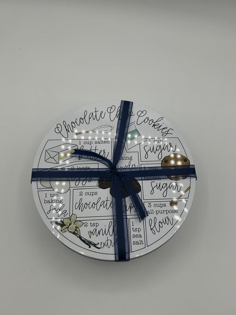 A beautifully wrapped Purim gift tin featuring a stylish chocolate chip cookie recipe design on the lid, secured with a navy blue ribbon. The tin contains baking essentials and treats, making it a perfect Mishloach Manos gift for Purim.