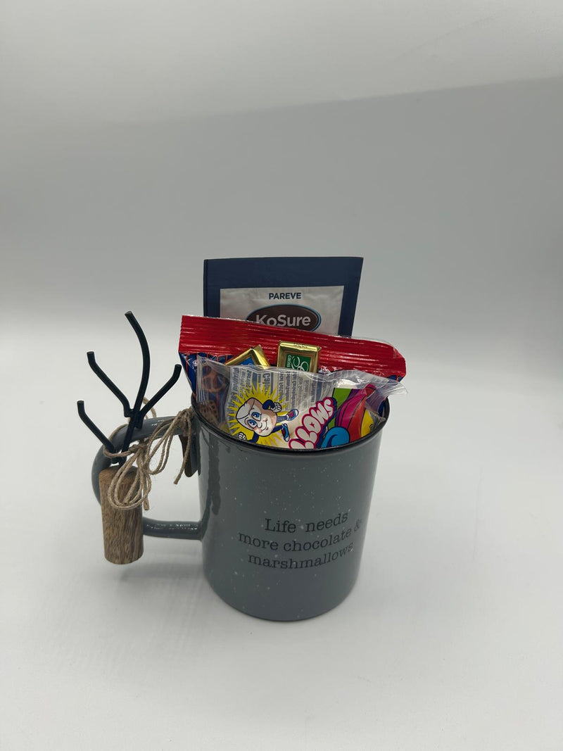 A close-up of the unwrapped Shalach Manos hot chocolate set, arranged on a white surface. The dark gray enamel mug with a roasting fork tied to the handle is accompanied by a pack of hot cocoa mix, mini marshmallows, pretzels, and two Alprose Chocolate Napolitains, making it the perfect winter treat-themed Purim gift.