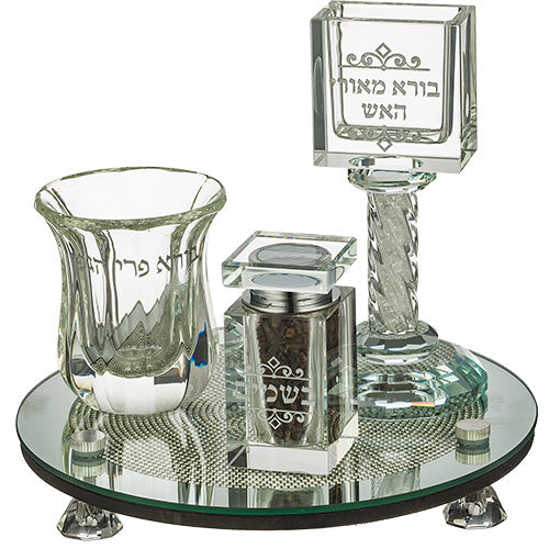 A beautifully designed clear crystal Havdalah set on a round mirror base with silver accents. The set includes a Kiddush cup, a square spice box, and a tall candle holder, all elegantly engraved with Hebrew text.