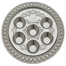 A round, silver-colored plastic Seder plate with an ornate embossed border, six labeled indentations for Seder foods, and a central cup for bitter herbs.