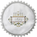 A round, white satin matzah cover with intricate embroidery featuring a pitcher, glasses, and the Hebrew word for Passover, bordered by a large, ornate silver fringe.