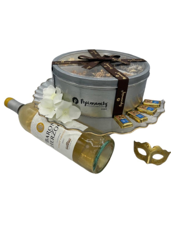 A luxurious Shalach Manos gift set featuring a Popinsanity deluxe tri-tin of gourmet popcorn, a bottle of Baron Herzog Pinot Grigio, and Alprose Napolitain chocolates, all arranged elegantly on a gold-rimmed reversible chip & dip/cake stand. A decorative gold mask and delicate white flowers add a festive Purim touch.