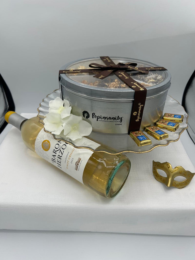 A beautifully wrapped Shalach Manos gift set, presented on a white gift box. The set includes a Popinsanity deluxe tri-tin of artisanal popcorn, a bottle of Baron Herzog Pinot Grigio, and Alprose Napolitain chocolates. The arrangement sits on an elegant gold-rimmed serving stand, with a gold masquerade mask placed nearby for a festive Purim aesthetic.