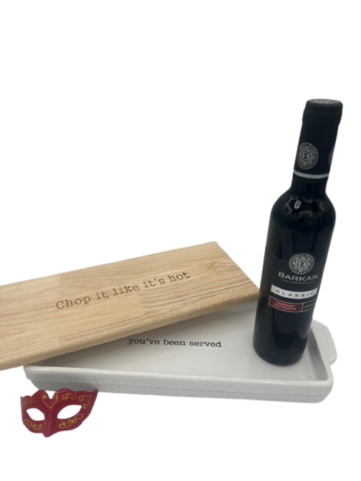 A stylish Shalach Manos gift set featuring a Mud Pie rectangular baking dish with a wooden lid engraved with ‘Chop it like it’s hot.’ A bottle of Barkan Classic Cabernet Sauvignon rests on top, accompanied by a decorative red and gold Purim mask.