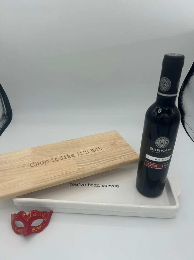 A close-up of the Shalach Manos set, with the Barkan Classic Cabernet Sauvignon placed on top of the Mud Pie wooden cutting board lid, which has the phrase ‘Chop it like it’s hot’ engraved. The baking dish below features ‘You’ve been served’ for a playful touch, with a decorative Purim mask beside it.