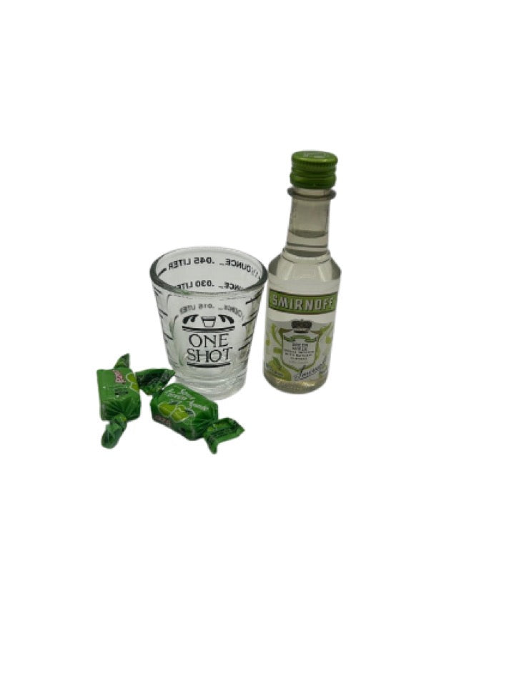 A mini bottle of Smirnoff Green Apple Vodka (50ml), a Fox Run 1.5 oz Measuring Shot Glass, and two sour green apple candies arranged on a clean white background.